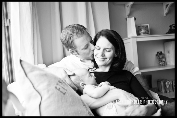 Portrait of family sitting on a couch with the father kissing his wife as she smiles, eyes closed, with their baby in her arms.