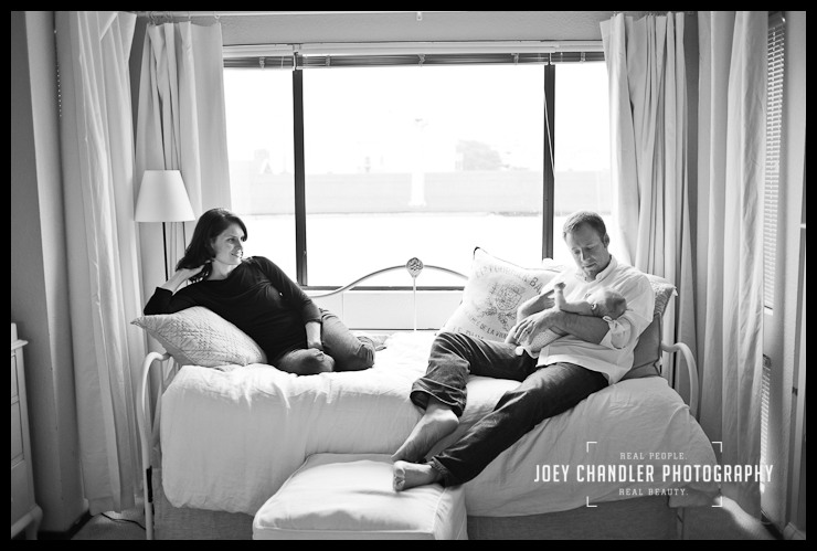 Lounging with the baby in their San Francisco home Black and white photo of a family sitting on a day bed. The mother is looking on from one side of the bed as her husband cradles their baby on the other side.