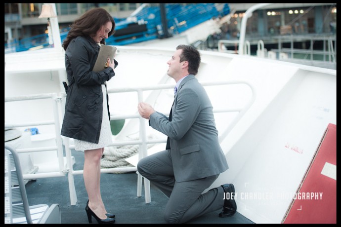 Man on one knee on the deck of a San Francisco ferry boat with a ring in his hand, looking up at a woman who has a look of surprise and excitement on her face.